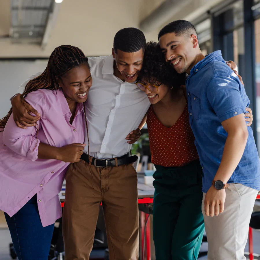 Cuatro colegas sonrientes y diversos se abrazan en un ambiente de oficina, mostrando trabajo en equipo y felicidad.