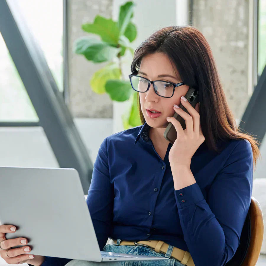 Mujer con gafas hablando por teléfono y usando una computadora portátil.