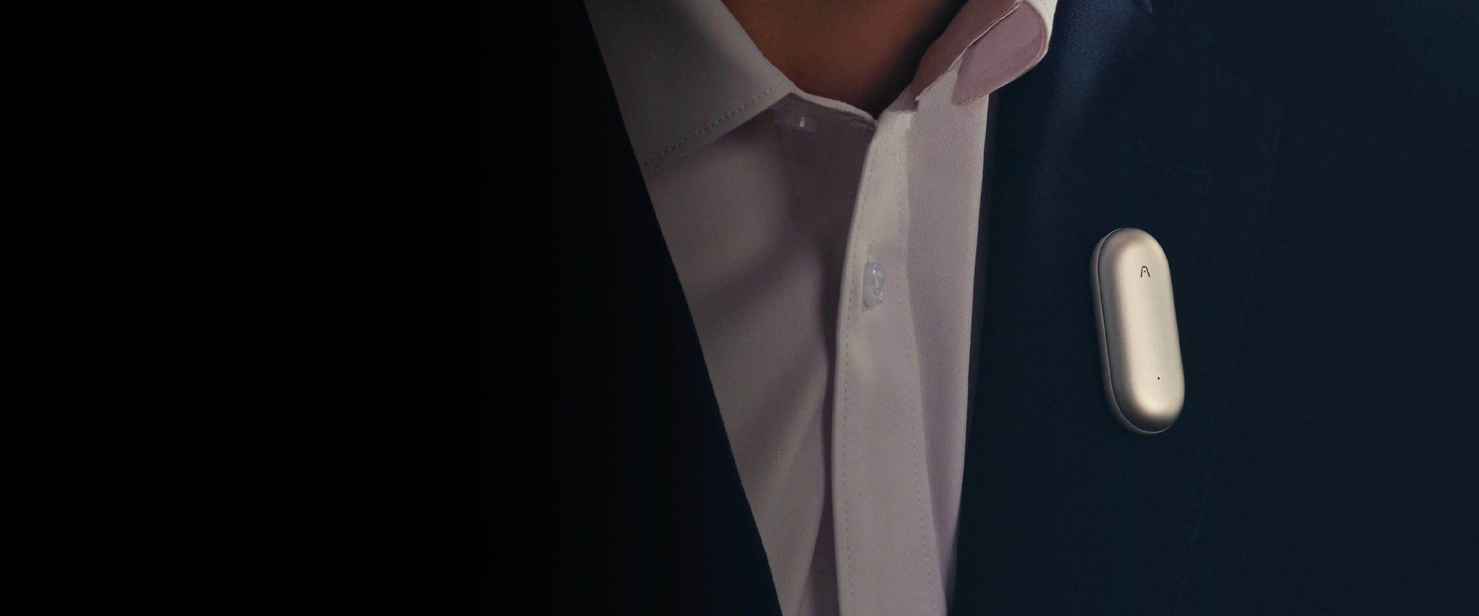Plaud NotePin plateado claro sujetado a una chaqueta oscura junto a una camisa blanca.