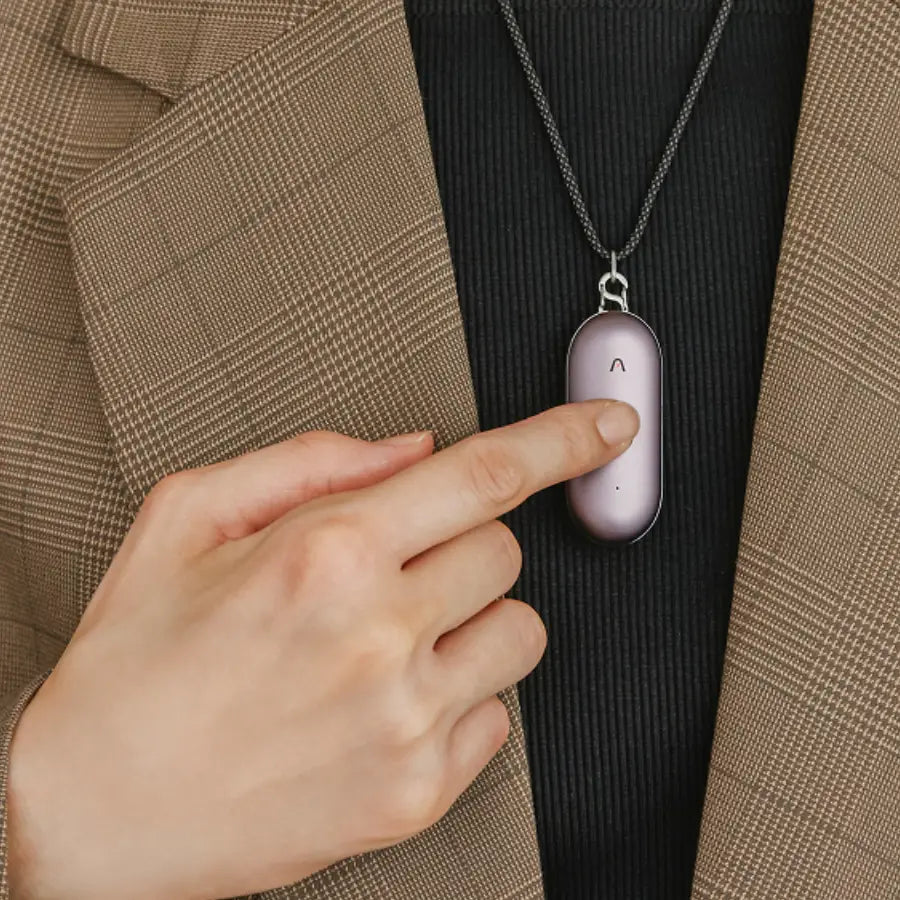 Mano presionando el Plaud NotePin lavanda, un grabador de voz AI, colgado de un collar sobre un blazer.