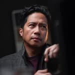 Hombre asiático con chaqueta oscura y camisa roja, mirando pensativo hacia arriba.