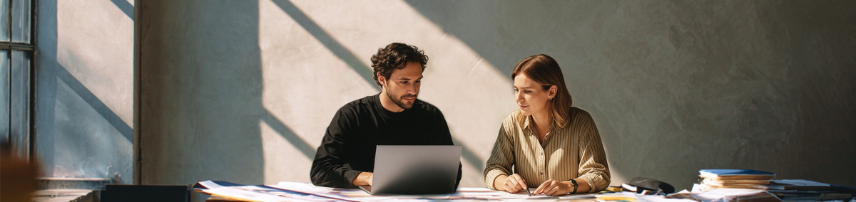 Dos personas colaborando en una laptop y documentos en una oficina iluminada por el sol.