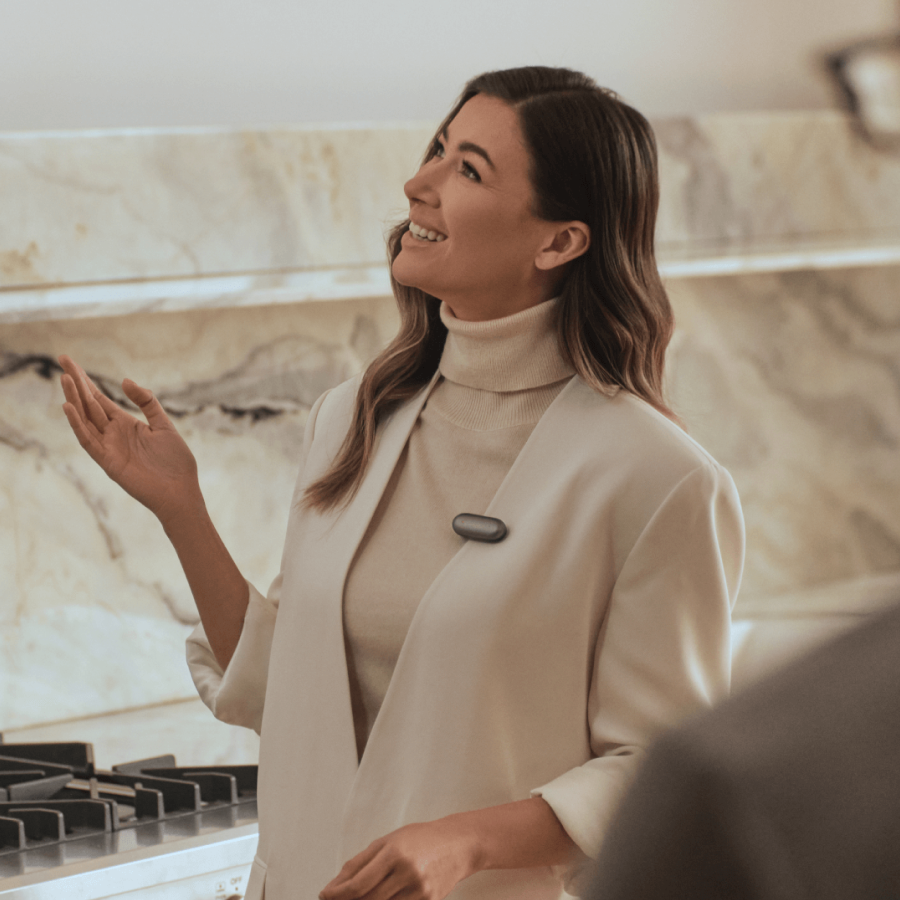 Mujer sonriente con un Plaud NotePin gris oscuro en su chaqueta, gesticulando en un interior moderno.