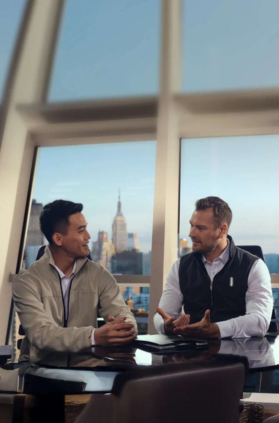 Dos hombres en una reunión de negocios, uno con un Plaud NotePin blanco, con el horizonte de la ciudad de Nueva York de fondo