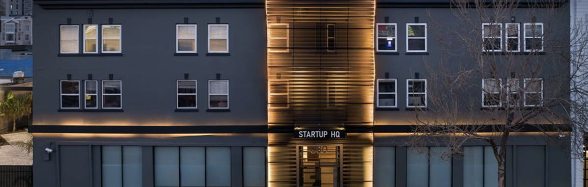 Edificio moderno gris oscuro con luces cálidas en la entrada y ventanas, mostrando el letrero 'STARTUP HQ'.