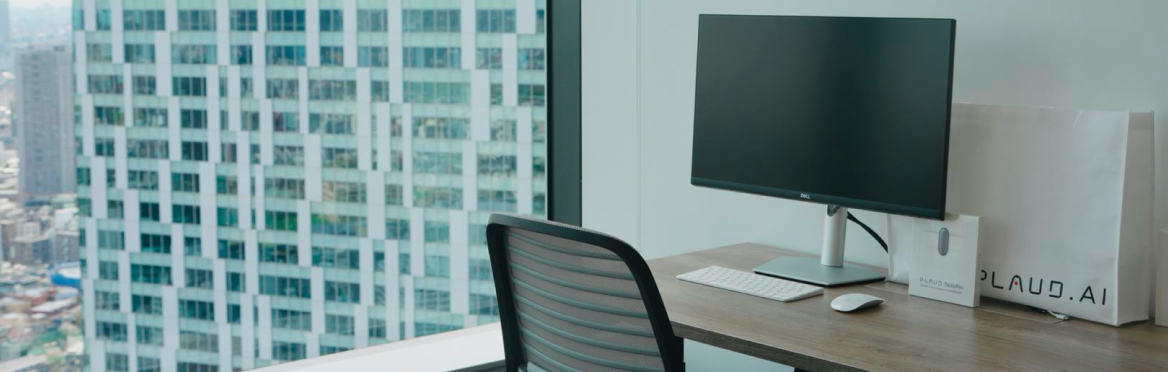 Escritorio moderno con monitor, teclado y bolsas Plaud.AI, incluyendo una Plaud Note, frente a una ventana con vista urbana.