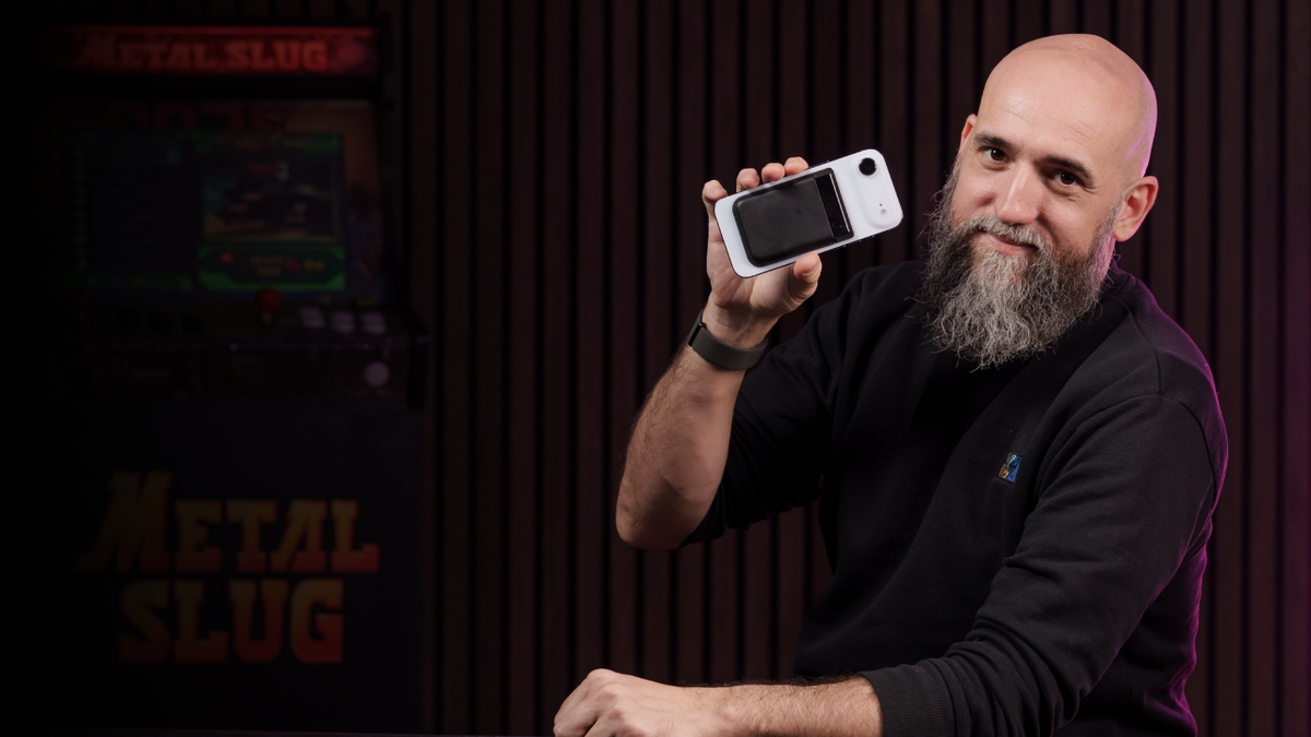 Hombre sonriente con barba sostiene un Plaud Note Pro plateado. Al fondo, una máquina arcade Metal Slug.