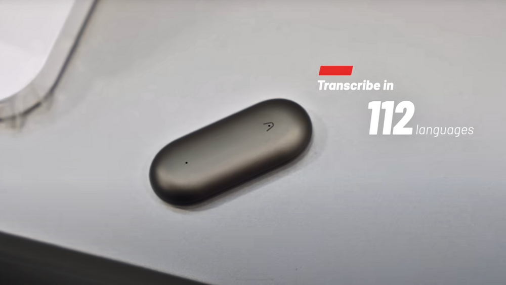 Plaud NotePin gris oscuro sobre una superficie clara, con texto "Transcribe en 112 idiomas".