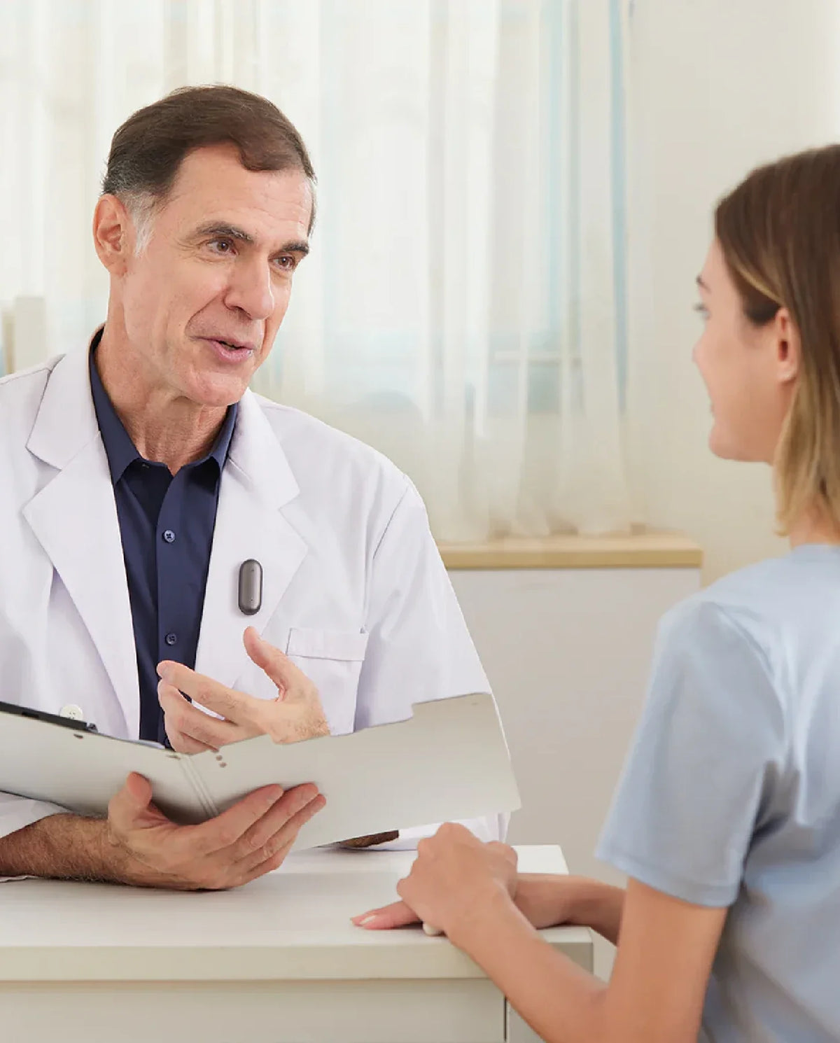 Doctor con un Plaud NotePin gris oscuro en su bata, hablando con una paciente en una consulta médica.