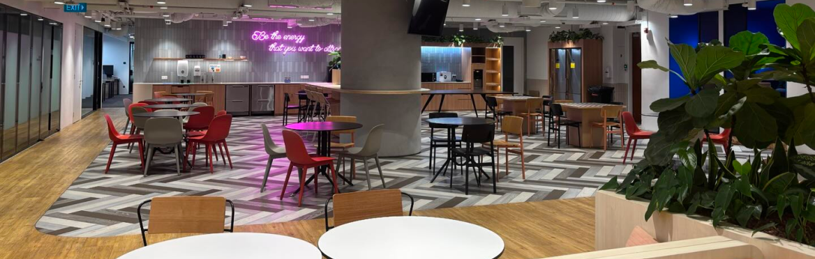 Moderna área común de oficina con mesas, sillas, cocina y plantas. Un letrero de neón dice "Be the energy that you want to